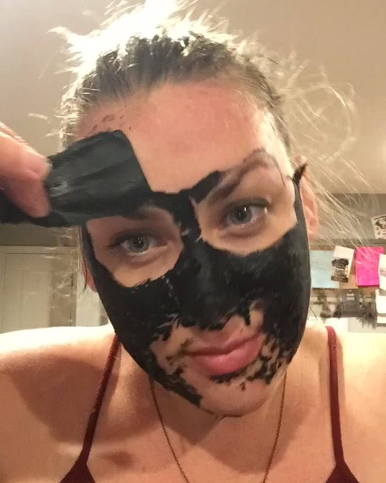 May include: A woman is applying a black charcoal face mask to her face. The mask is already applied to her forehead, nose, and chin.