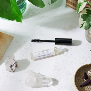 May include: A bottle of "Organics Eyelash Growth Serum" with a black mascara wand, set on a white surface. The image includes quartz crystals, a small decorative bowl, and green plants.