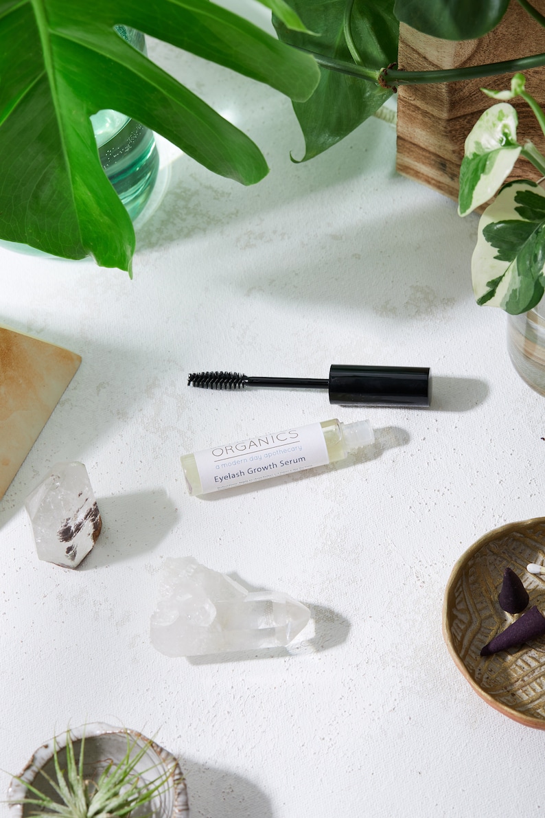 May include: A bottle of "Organics Eyelash Growth Serum" with a black mascara wand, set on a white surface. The image includes quartz crystals, a small decorative bowl, and green plants.