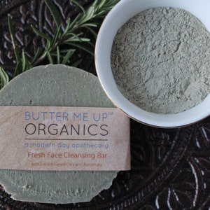 Puede incluir: Una barra de limpieza facial verde con una etiqueta marr&oacute;n que dice "BUTTER ME UP&trade; ORGANICS a modern day apothecary Fresh Face Cleansing Bar with French Green Clay and Rosemary". La barra est&aacute; sobre una superficie marr&oacute;n con una ramita de romero.