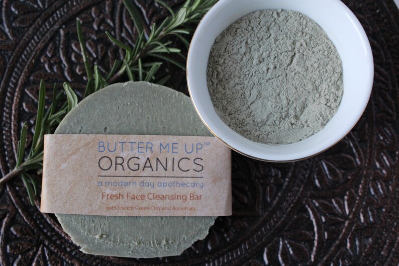 Puede incluir: Una barra de limpieza facial verde con una etiqueta marr&oacute;n que dice "BUTTER ME UP&trade; ORGANICS a modern day apothecary Fresh Face Cleansing Bar with French Green Clay and Rosemary". La barra est&aacute; sobre una superficie marr&oacute;n con una ramita de romero.