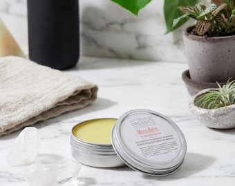 May include: A round silver metal tin containing yellow balm with a lid labeled Butter Me Up Organics Moon Balm. The lid features printed text detailing ingredients and usage instructions. The tin is placed on a white marble surface.