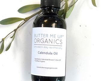 May include: A glass bottle of Calendula Oil from Butter Me Up Organics, a modern day apothecary. The bottle has a white label with the brand name, product name, and ingredients. The bottle is sitting on a white surface with a green branch.