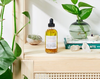 May include: A clear glass bottle of botanical body oil with a dropper top. The bottle is labelled "Butter Me Up Organics" and "Botanical Body Oil" with the scent "Mary Calenda". The bottle is sitting on a wooden shelf with a book and crystals.