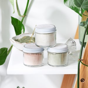 Op de afbeelding: Drie kleine glazen potjes met witte deksels die verschillende gekleurde poeders bevatten. De potjes zijn in een driehoekige vorm op een wit rek geplaatst. De potjes zijn gelabeld met "Illuminating Mask", "Rose Clay Mask" en "Green Clay Mask".