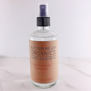 Könnte beinhalten: Eine durchsichtige Glasflasche mit schwarzer Sprühdüse und einem braunen Etikett mit der Aufschrift "BUTTER ME UP ORGANICS a modern day apothecary Fabric & Room Refresher Lavender 8 fl. oz / 238 ml"