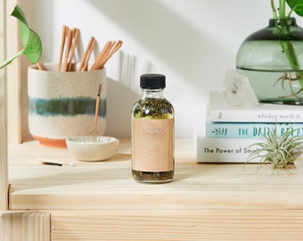 Peut inclure: Une bouteille en verre transparent avec un bouchon noir remplie d'un liquide vert clair et d'herbes séchées. La bouteille est étiquetée "ELEMENTAL ORGANICS" et "Bath Oil".