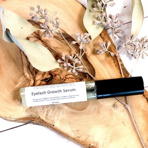 May include: A clear glass roller bottle of eyelash growth serum with a black cap, resting on a wooden surface. The label reads "Eyelash Growth Serum" and "10 ml". Dried foliage and a wooden slab are in the background.