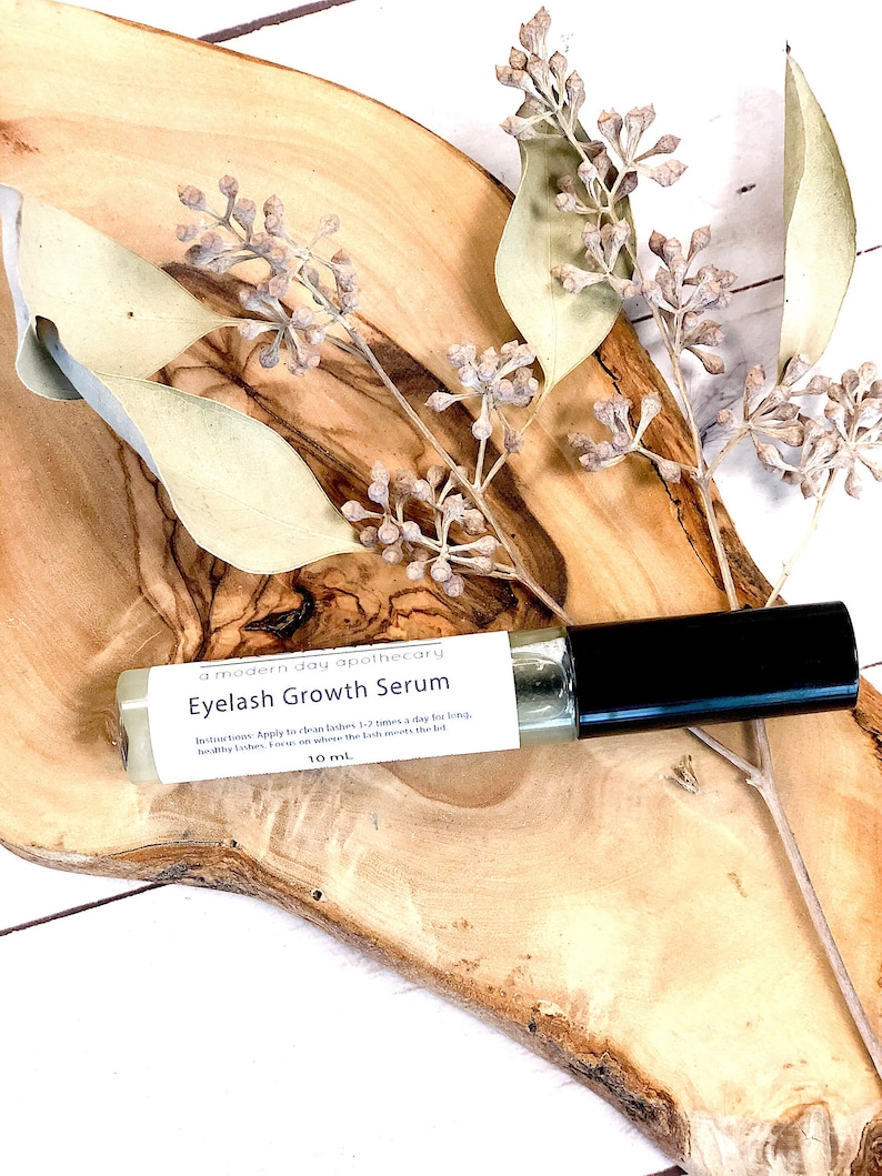 May include: A clear glass roller bottle of eyelash growth serum with a black cap, resting on a wooden surface. The label reads "Eyelash Growth Serum" and "10 ml". Dried foliage and a wooden slab are in the background.