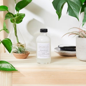 May include: A clear glass bottle with a black cap and a white label that reads "Butter Me Up Organics" and "Makeup Remover". The bottle is sitting on a wooden surface with green plants in the background.