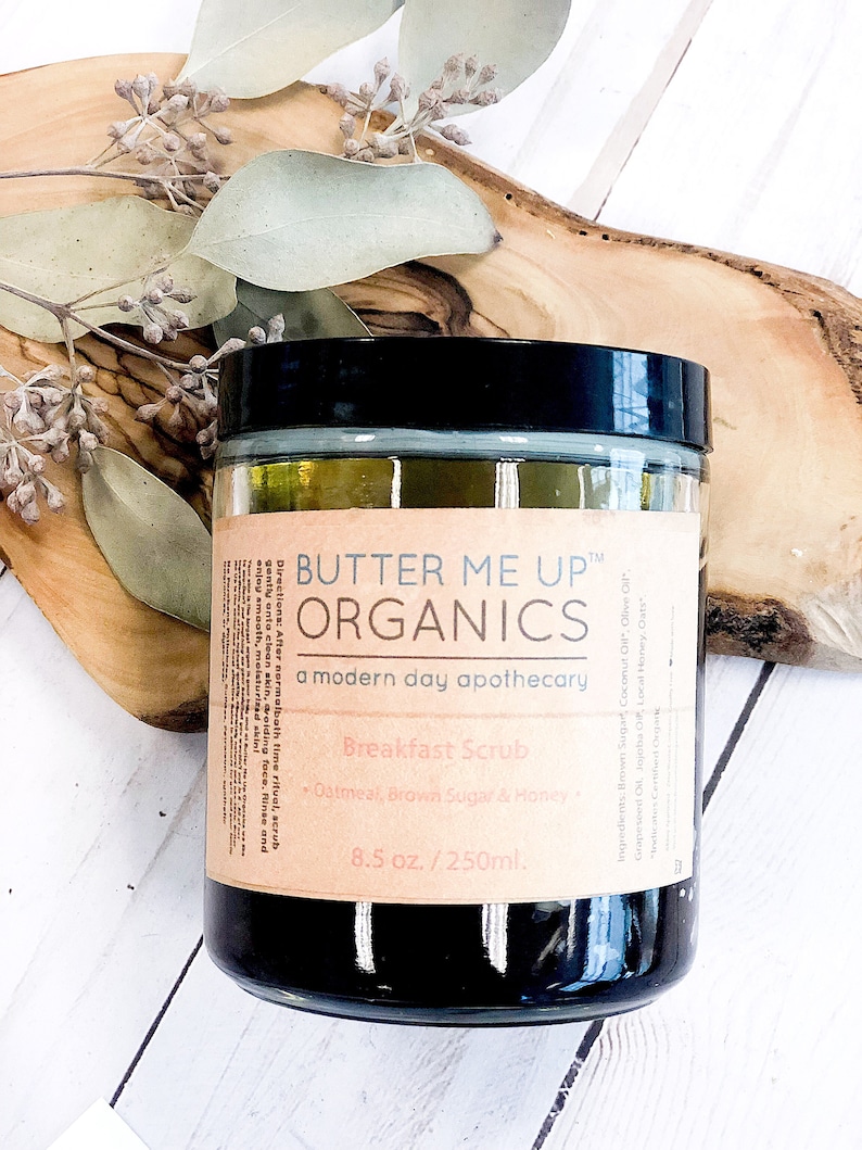 May include: A jar of "Butter Me Up Organics" Breakfast Scrub, 8.5 oz. size, with a black lid and a brown label. The label includes the ingredients: Brown Sugar, Coconut Oil, Olive Oil, Grapeseed Oil, Jojoba Oil, Local Honey, Oats. The jar is on a wooden surface.