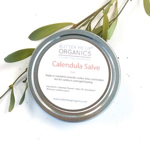 May include: A silver tin of Calendula Salve by Butter Me Up Organics, a modern day apothecary. The label is white with black text and says "Calendula Salve" and "2 oz". The ingredients are listed as Calendula Flowers, Olive Oil, and Shea Butter.