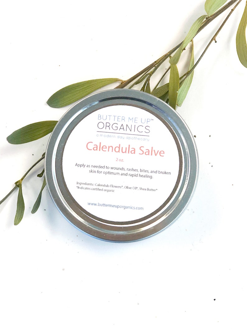 May include: A silver tin of Calendula Salve by Butter Me Up Organics, a modern day apothecary. The label is white with black text and says "Calendula Salve" and "2 oz". The ingredients are listed as Calendula Flowers, Olive Oil, and Shea Butter.