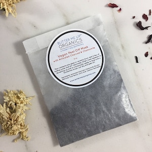 May include: A white paper pouch with a black powder inside. The pouch has a sticker on the front that says "Butter Me Up Organics" and "Vegan Peel Off Mask with Activated Charcoal & Chamomile".