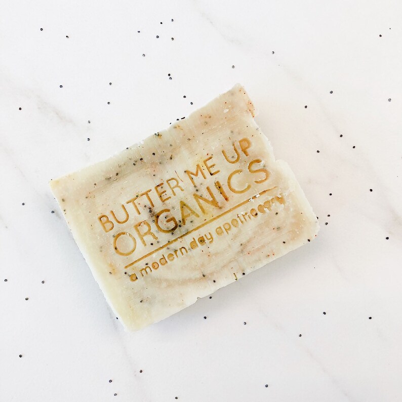 K&ouml;nnte beinhalten: Eine wei&szlig;e Seifenstange mit einem goldenen Logo, auf dem "Butter Me Up Organics a modern day apothecary" steht.