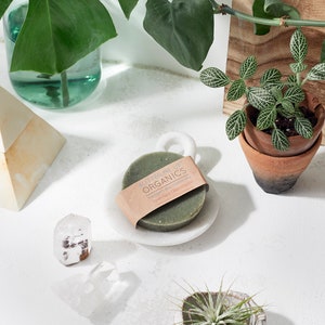 Puede incluir: Una barra de jab&oacute;n verde redonda con el texto "Butter Me Up Organics" y "Vegan Face Cleansing Bar" en un jabonera de m&aacute;rmol blanco. El jab&oacute;n est&aacute; rodeado de plantas y cristales.