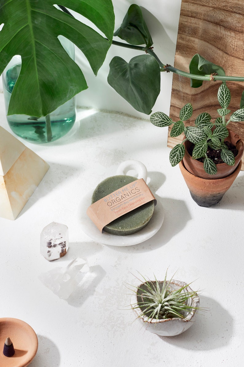 Puede incluir: Una barra de jab&oacute;n verde redonda con el texto "Butter Me Up Organics" y "Vegan Face Cleansing Bar" en un jabonera de m&aacute;rmol blanco. El jab&oacute;n est&aacute; rodeado de plantas y cristales.