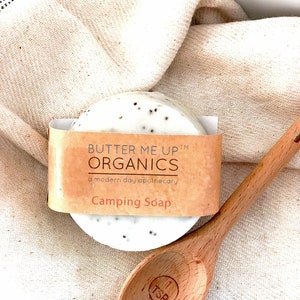 Könnte beinhalten: Eine runde, weiße Seife mit schwarzen Punkten, verpackt in braunes Papier mit dem Text "Butter Me Up™ Organics a modern day apothecary Camping Soap". Die Seife liegt auf einer weißen Stoffoberfläche neben einem Holzlöffel.