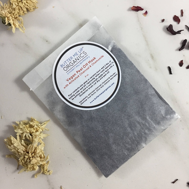 May include: A white paper pouch with a black powder inside. The pouch has a label that says "Butter Me Up Organics" and "Vegan Peel Off Mask with Activated Charcoal & Chamomile".