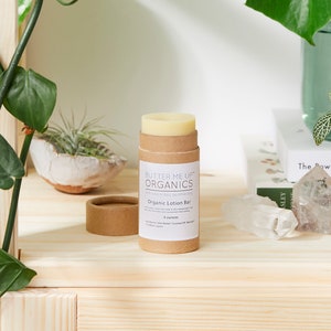 Puede incluir: Un tubo de cartón marrón con una etiqueta blanca que dice "Butter Me Up Organics" y "Organic Lotion Bar". El tubo contiene una barra de loción sólida blanca. El tubo está sentado en un estante de madera con una planta verde en el fondo.