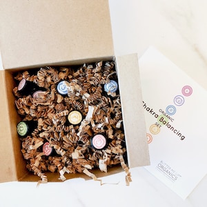 May include: A brown cardboard box with its lid open, revealing a set of small glass bottles with colourful labels. The bottles are nestled in brown packing paper. The box is labelled "Organic Chakra Balancing Set" and "Butter Me Up Organics".