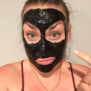 May include: A person with a black charcoal face mask on their face. The mask covers their entire face, including their eyes, nose, and mouth. The person is looking at the camera and has a slight smile on their face.
