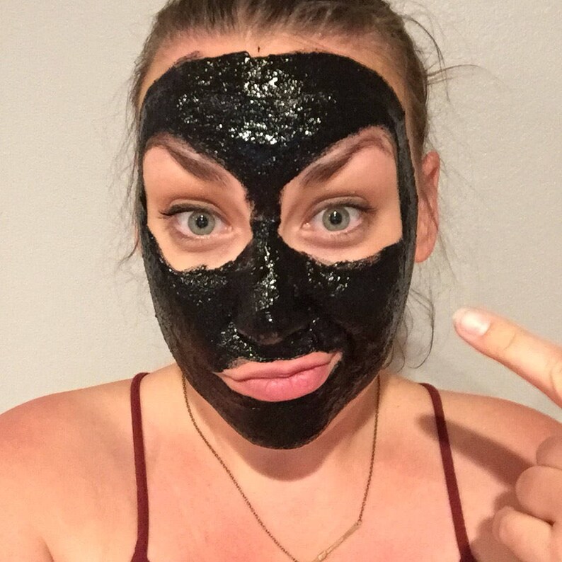 May include: A person with a black charcoal face mask on their face. The mask covers their entire face, including their eyes, nose, and mouth. The person is looking at the camera and has a slight smile on their face.
