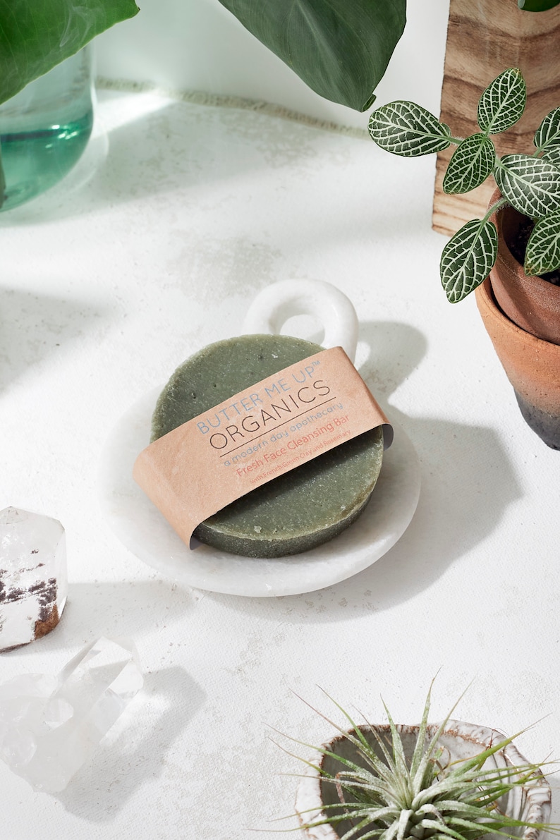 Puede incluir: Una barra de limpieza facial verde con una etiqueta de papel marr&oacute;n que dice "Butter Me Up Organics" y "Fresh Face Cleansing Bar". La barra est&aacute; en un plato de m&aacute;rmol blanco con un asa.