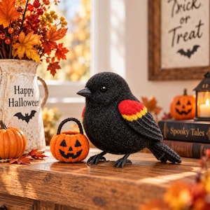 Könnte beinhalten: Eine schwarze Filzvogel-Figur mit einem roten und gelben Flügelabzeichen, platziert neben einem kleinen orangefarbenen gehäkelten Kürbiskorb mit einem schwarzen Jack-o'-Lantern-Gesicht. Der Vogel hat einen dunklen Schnabel und einen strukturierten schwarzen Körper.