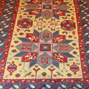 Puede incluir: Una alfombra rectangular con un campo de color crema y un gran motivo de estrella central en azul y rojo. El diseño incluye patrones geométricos, figuras de animales estilizadas y elementos florales. El borde es rojo oscuro con detalles azules.