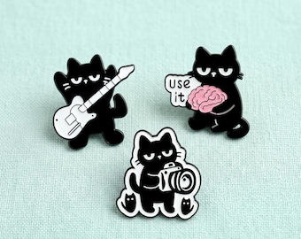May include: Three black cat enamel pins. One cat holds a white electric guitar, another holds a pink brain with a speech bubble reading USE IT, and the third cat holds a black and white camera with two smaller cat silhouettes at its feet.