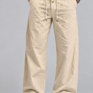 Op de afbeelding: Beige broek met wijde pijpen, een knoopsluiting met drie zichtbare bruine knopen en een trekkoord in de taille. De broek heeft een textuur in de stof en zijzakken, gecombineerd met een wit overhemd en witte sneakers.
