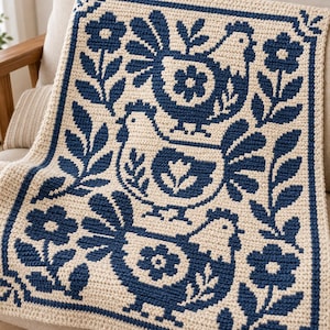 Op de afbeelding: Een rechthoekige gehaakte deken met een crèmekleurige basis en een donkerblauw patroon. Het ontwerp toont drie gestapelde kippen omringd door bloem- en bladmotieven, allemaal omsloten door een donkerblauwe rand met accenten in de hoeken.