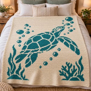 May include: A cream-colored crochet blanket featuring a large teal sea turtle swimming upward. The design includes teal bubbles rising from the turtle, along with teal seaweed and coral motifs at the bottom corners of the rectangular textile piece.