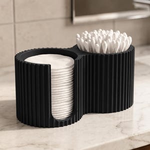 Puede incluir: Un organizador de baño negro de dos compartimentos con una textura estriada vertical. Un lado contiene una pila de discos de algodón blancos redondos, mientras que el otro lado contiene un grupo de bastoncillos de algodón blancos en forma de ocho.