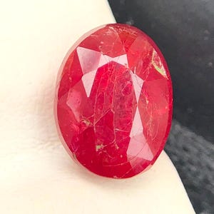Può includere: Una gemma di forma ovale di un rosso vibrante con una superficie sfaccettata. La pietra mostra una profonda tonalità cremisi e presenta inclusioni interne visibili e fratture naturali in tutta la sua struttura, riflettendo la luce sulla superficie lucida.