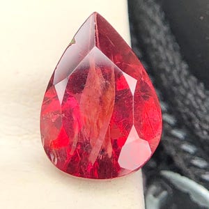 Può includere: Una gemma sfaccettata a forma di pera di colore rosso intenso. La pietra presenta una parte superiore appuntita e una base arrotondata, con molteplici sfaccettature geometriche sulla superficie che riflettono la luce, rivelando texture interne.