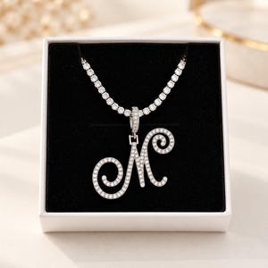 Può includere: Una collana color argento con un ciondolo a forma di lettera M corsiva incastonato di pietre chiare. Il ciondolo pende da una catena tennis abbinata, entrambi decorati con pietre scintillanti, presentati in una scatola bianca con interno nero.