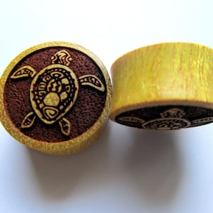May include: A pair of wooden ear gauges with a carved turtle design. The gauges are yellow and brown with a dark brown background.