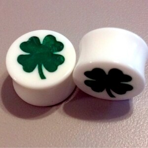 May include: Two white ear gauges with a four-leaf clover design. One gauge has a green clover, the other has a black clover.
