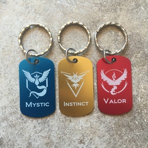 Op de afbeelding: Drie sleutelhangers met metalen ringen en rechthoekige labels. De labels zijn blauw, goud en rood. Elk label heeft een wit Pokémon-logo en de woorden "Mystic", "Instinct" en "Valor".