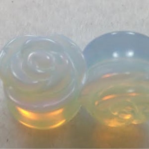 Puede incluir: Par de tapones de rosa de ópalo iridiscente. Los tapones son de color azul claro con un patrón de espiral.