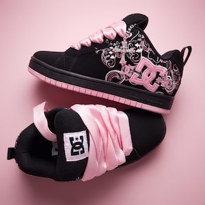 May include: A pair of black and pink DC sneakers featuring wide satin ribbon laces, a prominent pink DC logo on the side, and a decorative silver-toned cross charm attached to the eyelets. The shoes have a black base with pink floral patterns.
