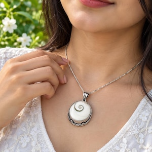 Puede incluir: Un collar con colgante redondo que presenta una concha de ojo de Shiva blanca con un patrón de espiral natural, engastada en un marco de metal plateado decorativo con un borde de cuerda trenzada, suspendido de una cadena de eslabones plateada.
