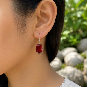 May include: A silver drop earring featuring a rectangular, faceted deep red gemstone held by four prongs. The earring hangs from a simple silver hook wire, showcasing a clear, polished finish on the stone and a metallic luster on the silver setting.