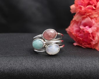 May include: A silver ring featuring three stacked bands, each topped with an oval-shaped gemstone. The stones include a dusty pink cabochon, a smooth light blue turquoise-colored stone, and a translucent white moonstone-like gem.