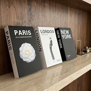 May include: Three decorative books featuring black and white covers. The first is black with white text reading PARIS CITY OF FASHION AND ROMANCE and a white flower. The second is white with LONDON OXFORD STREET text and a figure. The third is black with NEW YORK text.
