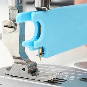 May include: A light blue plastic needle threader tool attached to a metal sewing machine presser foot and needle. The tool features a notched design that aligns with the gold-colored sewing machine needle to assist with threading the eye.
