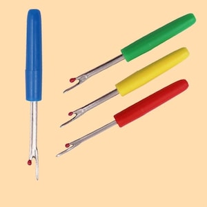 May include: Four seam rippers with plastic handles in blue, green, yellow, and red. Each tool features a metal shaft with a sharp, forked blade and a small red safety ball at the tip. The blue ripper is shown vertically, while the others are angled.