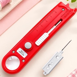May include: A bright red sewing tool featuring a built-in magnifying glass, a sliding white button mechanism, and a ruler printed along the side with markings up to 5 inches. A separate white needle-like attachment is positioned next to the tool.